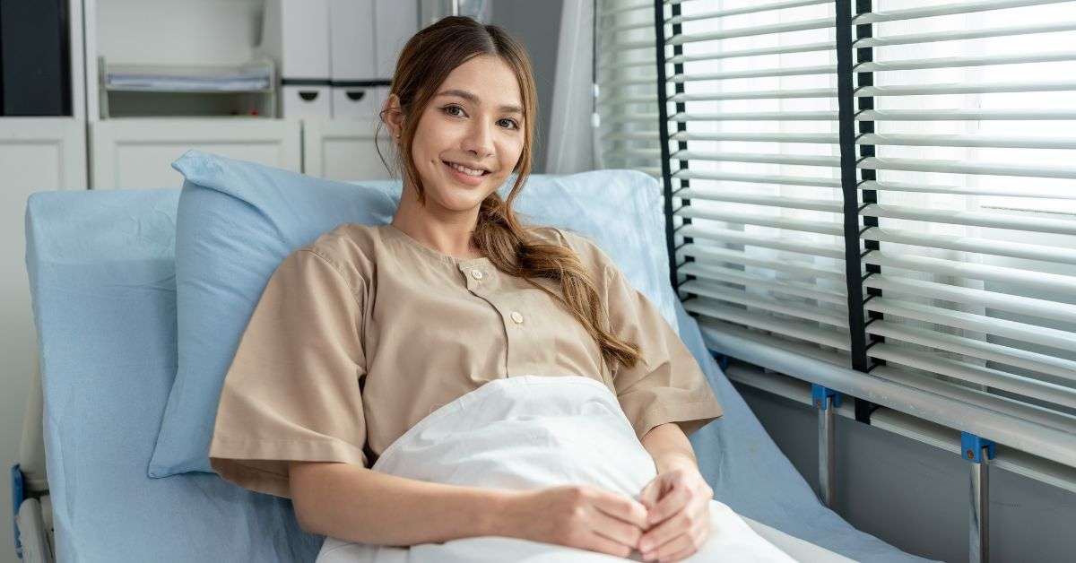A young woman smiles as she sits in an upright position in a home hospital bed next to a window with blinds.