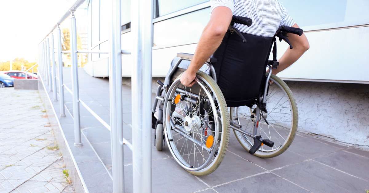 A wheelchair user using an exterior ADA wheelchair ramp. The ramp is steel with a low slope and a handrail.