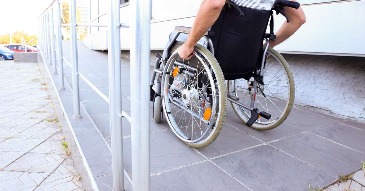 A person in a wheelchair rides their way up the ramp. The ramp is metal and the full-length of the sidewalk.