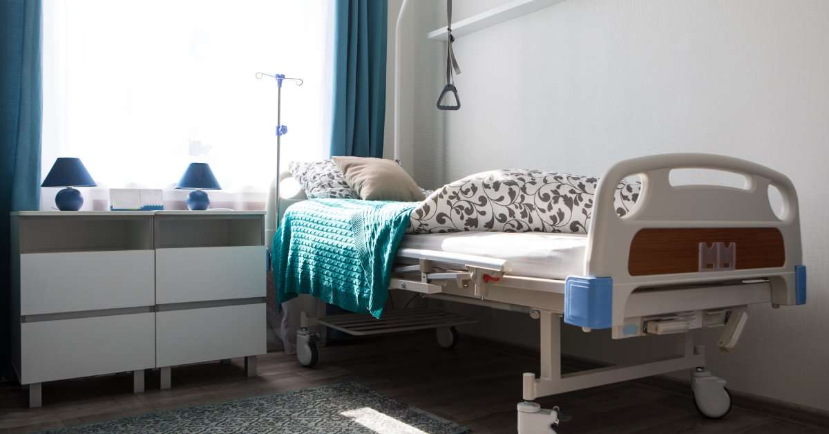 A hospital bed with bedsheets, blankets, and pillows sits in a residential room next to a rug and dresser.