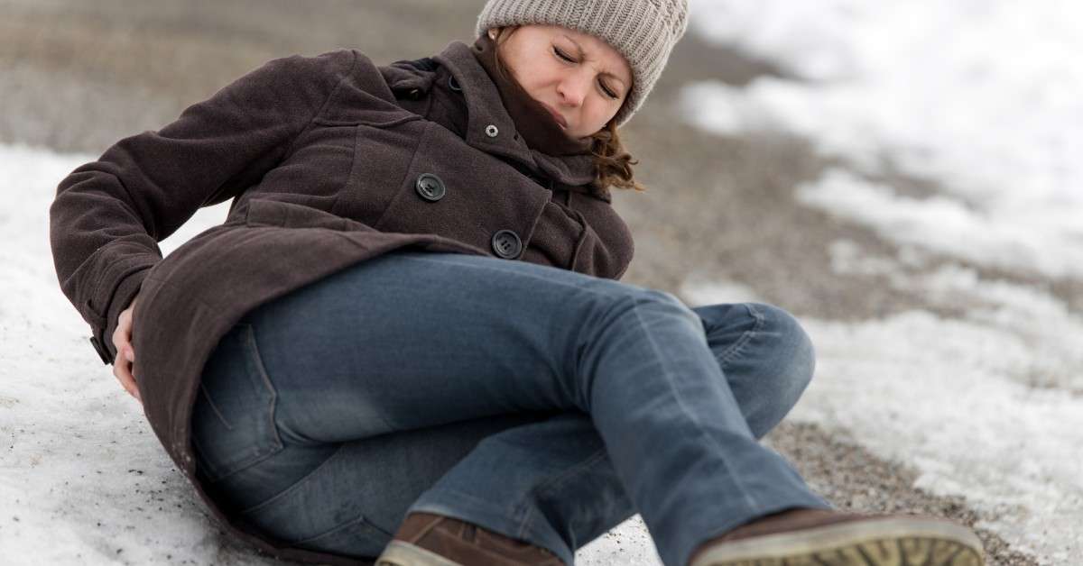 A woman lays on the ground after falling on the sidewalk. She's wearing a coat, winter hat, and boots, holding her back.
