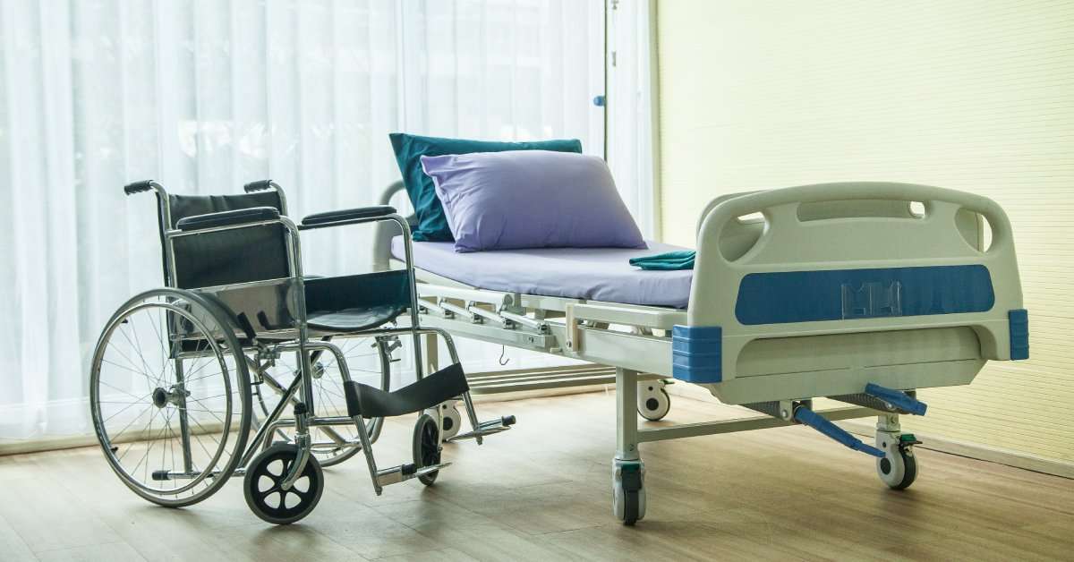 A wheelchair sits next to a hospital bed in the corner of a room with a large window in the background.