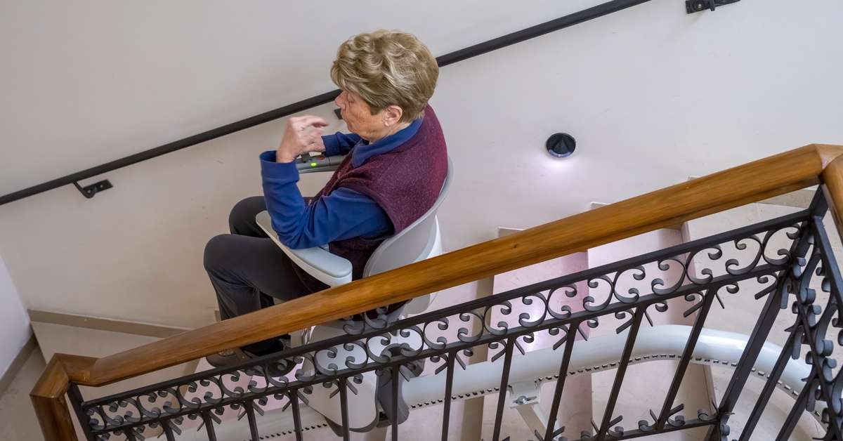 A senior woman rides her stairlift down a flight of stairs in her home. The handrail is wooden with black detailing.