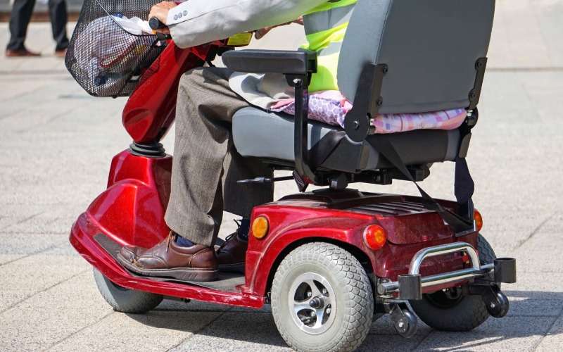 An aging adult rides a red electric wheelchair outdoors. They wear a high-vis safety vest and the basket has items.