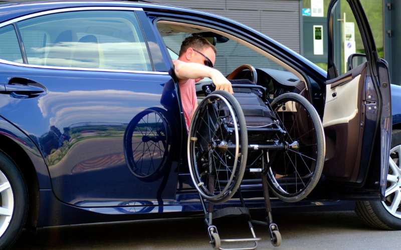 A male driver gets into a navy blue passenger vehicle as he lifts his wheelchair into the car with him.