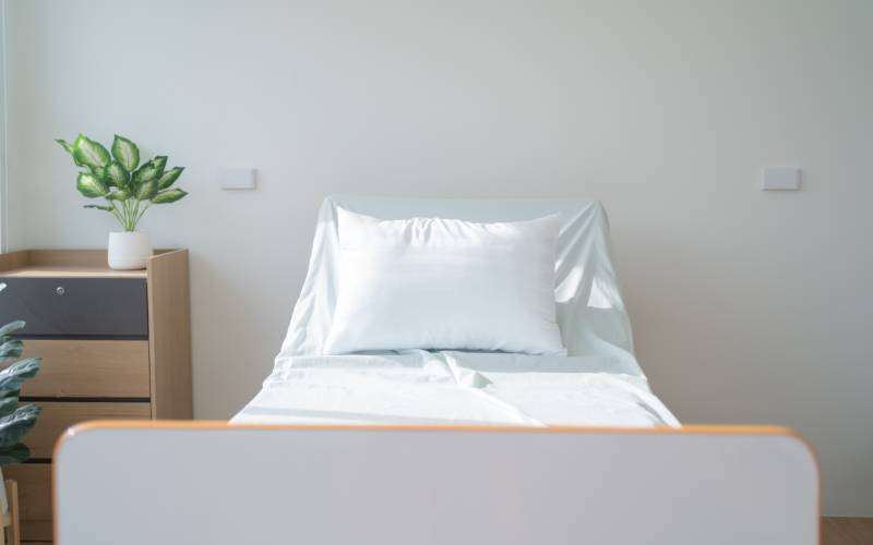 A home medical bed with white linens sits independently in a room. There is a nightstand to the left of the bed.
