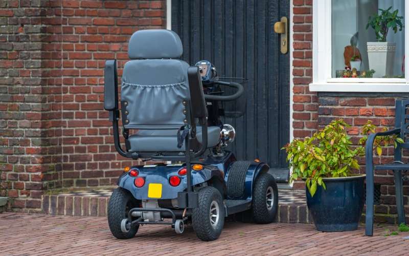 A mobility scooter parked outside a brick building. The building is residential and there's a plant and bench nearby.