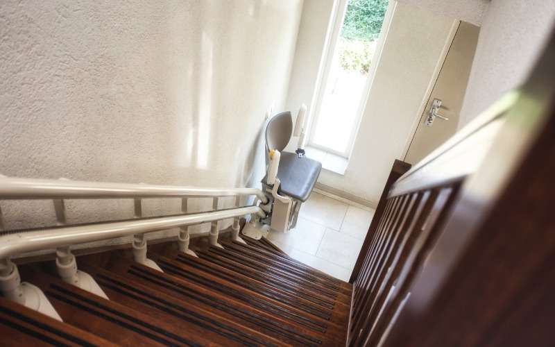 A stairlift at the very bottom of a flight of steps in a home. The stairs are hardwood and there's a window at the bottom.