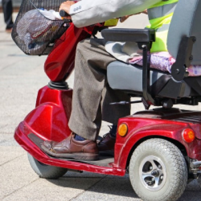 An aging adult rides a red electric wheelchair outdoors. They wear a high-vis safety vest and the basket has items.