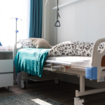 A hospital bed with bedsheets, blankets, and pillows sits in a residential room next to a rug and dresser.