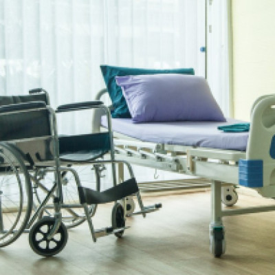 A wheelchair sits next to a hospital bed in the corner of a room with a large window in the background.