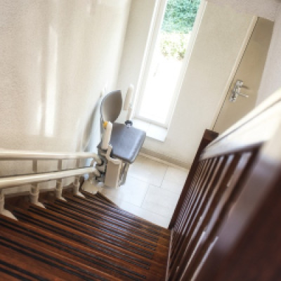 A stairlift at the very bottom of a flight of steps in a home. The stairs are hardwood and there