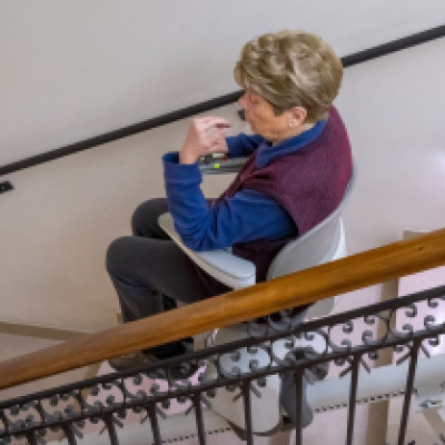 A senior woman rides her stairlift down a flight of stairs in her home. The handrail is wooden with black detailing.