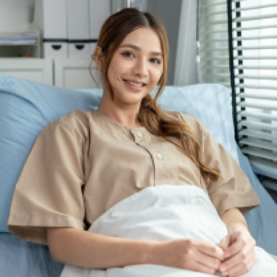 A young woman smiles as she sits in an upright position in a home hospital bed next to a window with blinds.