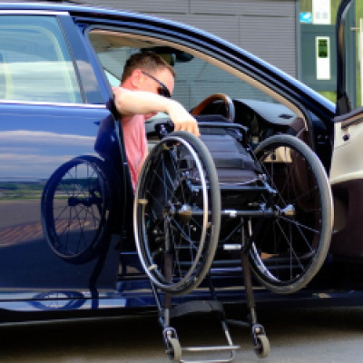 A male driver gets into a navy blue passenger vehicle as he lifts his wheelchair into the car with him.
