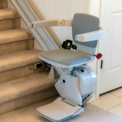 An electric chair at the bottom of a staircase life in a home. The stairs have beige-brown carpet and a wooden handrail.