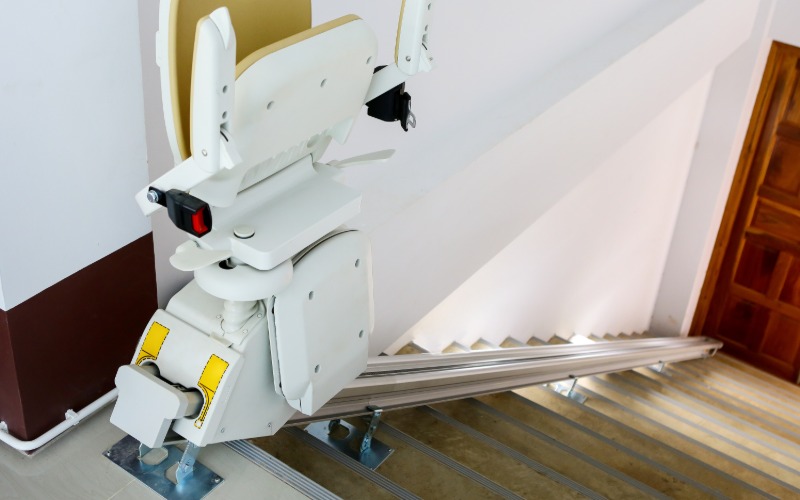 A stairlift seat at the top of the stairway not in use. The chair is folded up for safety and the stairway is lit up.