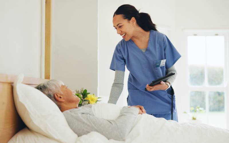 A home medical provider in blue scrubs stands bedside with a patient who lays in an adjustable hospital bed.