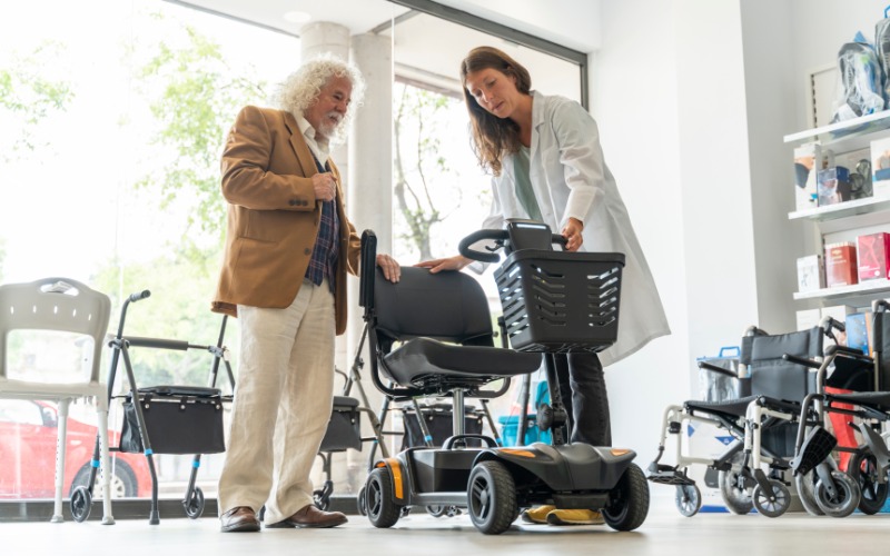An aging male stands in a medical supply store with his mobility scooter as a professional helps him out.