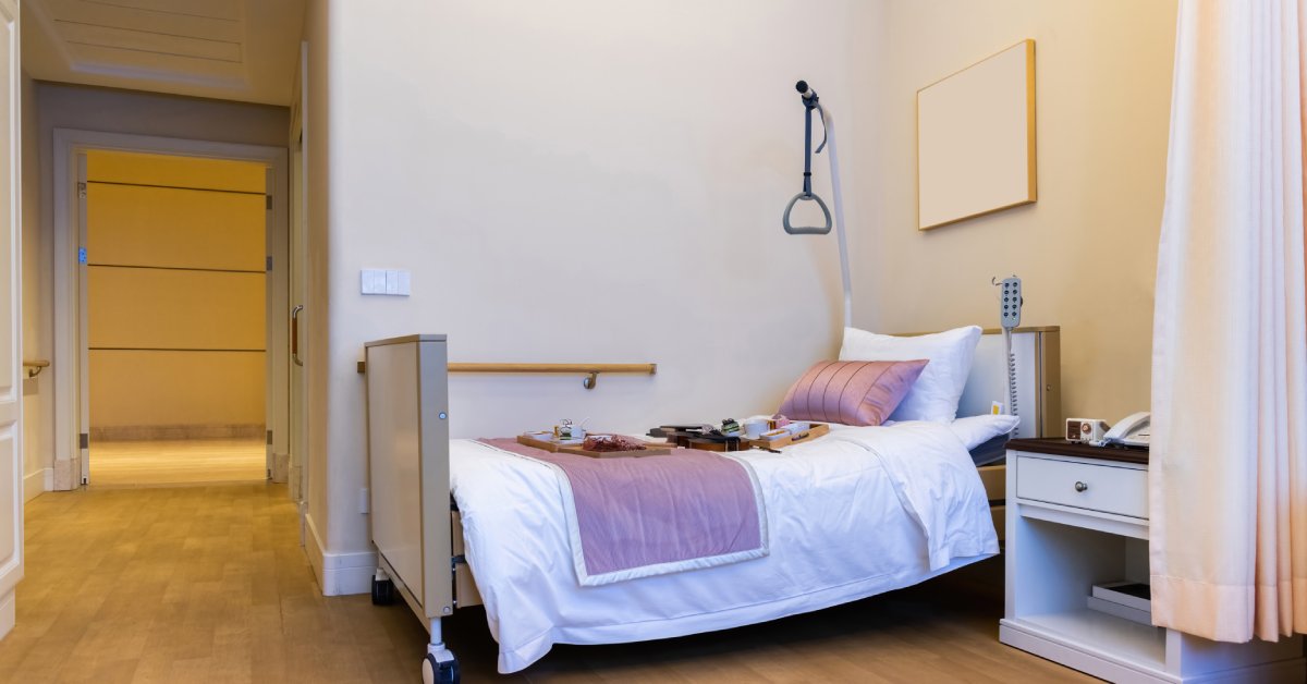 A large bedroom with an at-home hospital bed with a pull-up handle in the corner, sitting next to a bedside table.