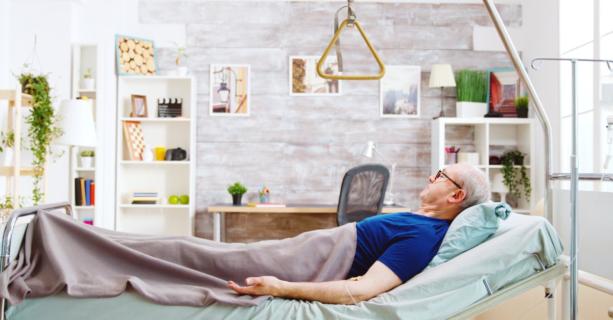 Closeup of an older man lying in an at-home hospital bed that has a pull-handle hanging above him for mobility.