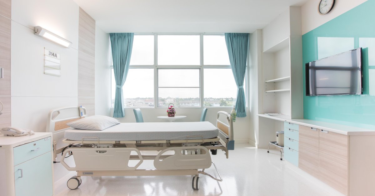 A hospital bed sits in a room with a TV on the opposite wall, cabinets, and a large window with blue curtains.