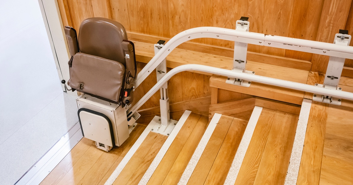 A brown and white electric stairlift chair with the seat folded up rests at the bottom of a short staircase.