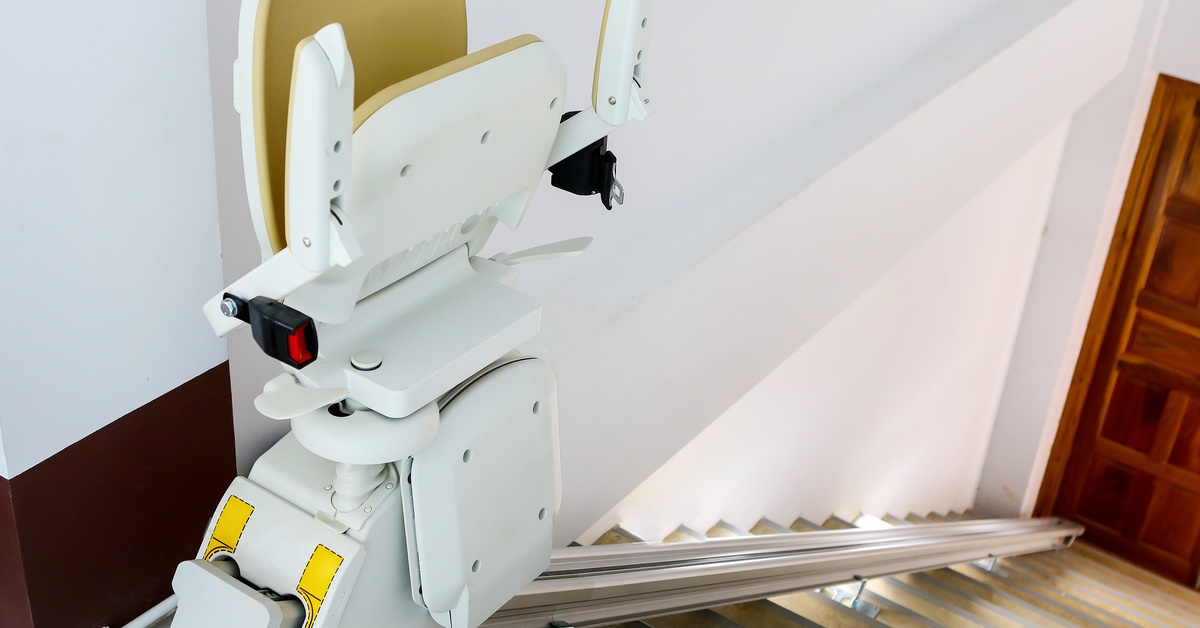 A yellow and white electric stairlift chair with the seat folded up is at the top of a steep wooden staircase.