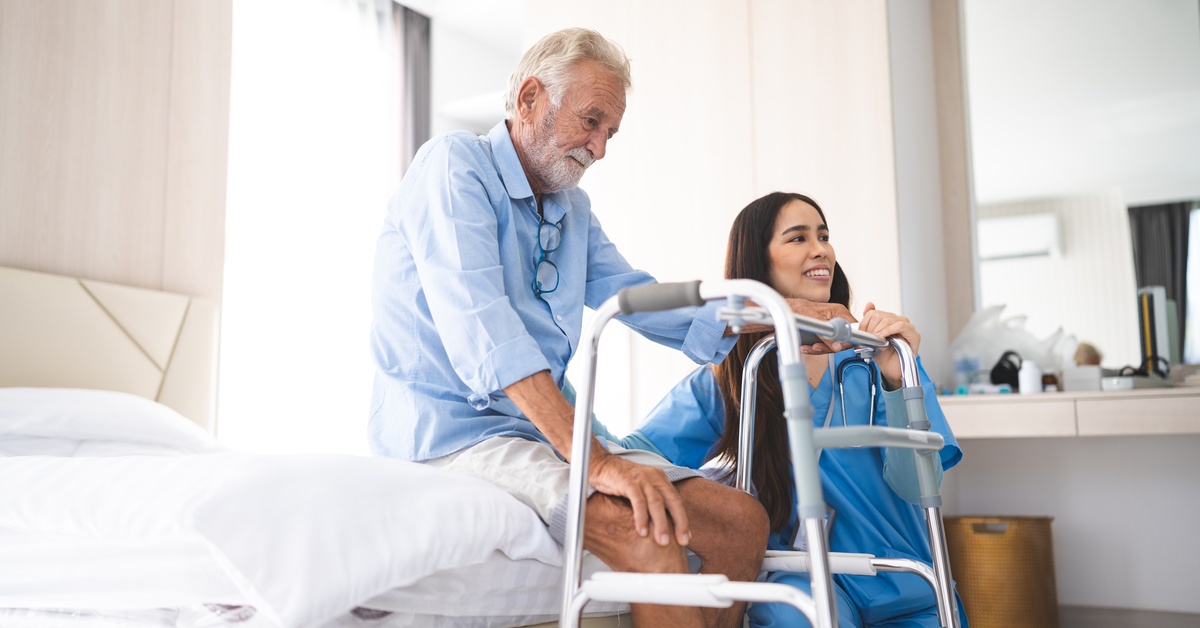 An elderly man sits on the edge of a bed and holds onto a walker mobility aid as a young female nurse assists him.