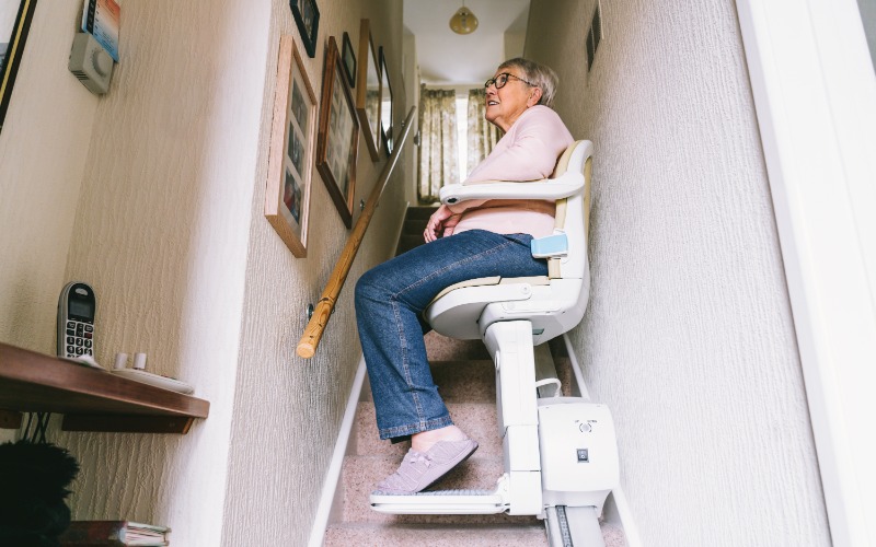 A senior woman at home rides an installed stairlift up the carpeted steps. She wears jeans and sneakers.