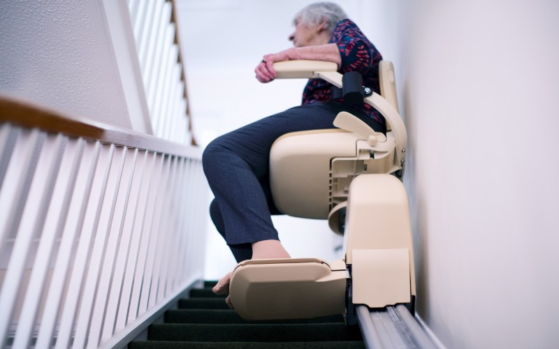 A senior person rides the stairlift in their home. The steps are black and the railing is white with a wood banister.