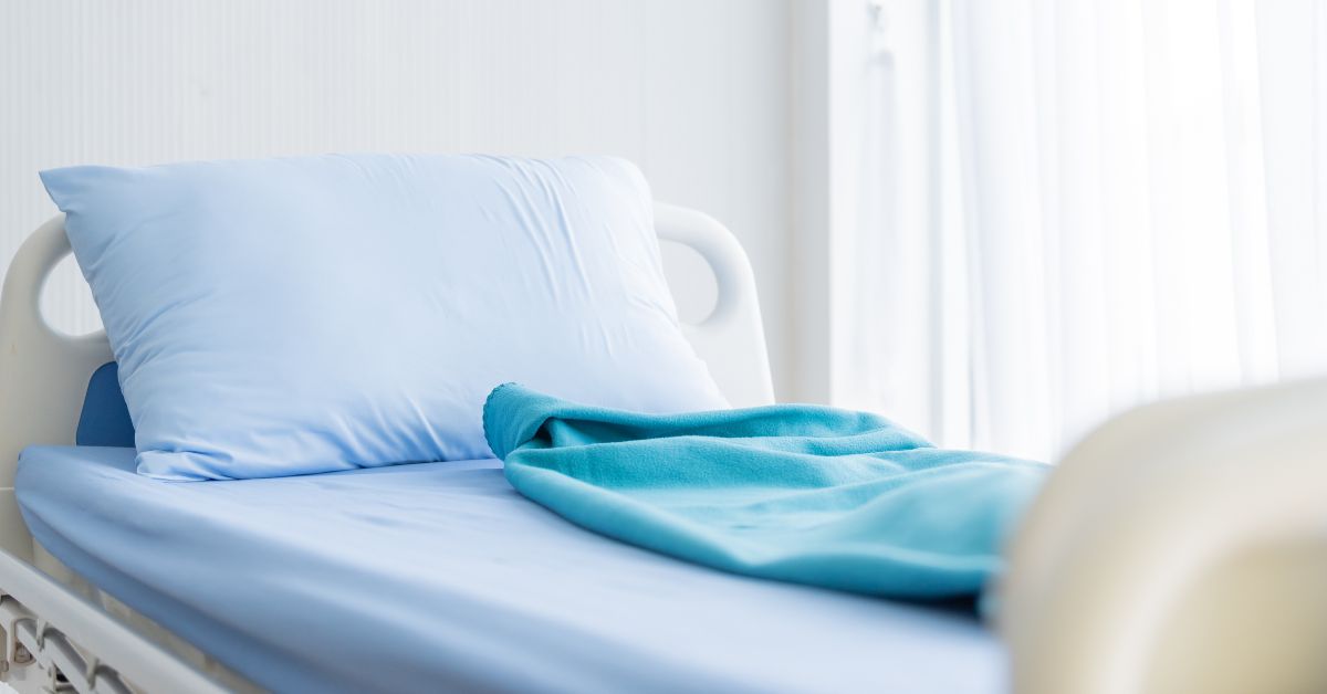 Close-up of a home hospital bed with a pillow, blue sheets, and a teal blanket sitting up against a white wall.