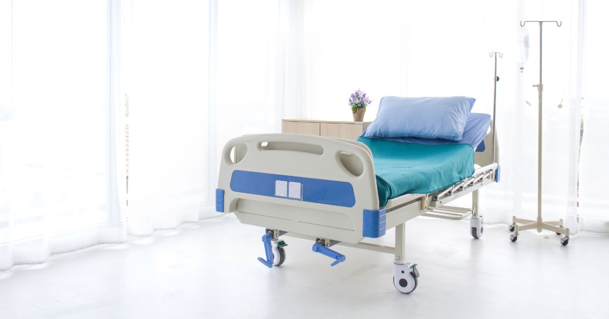A hospital bed with a pillow and blue sheets sits in a room next to an IV tree and a large window with white curtains.