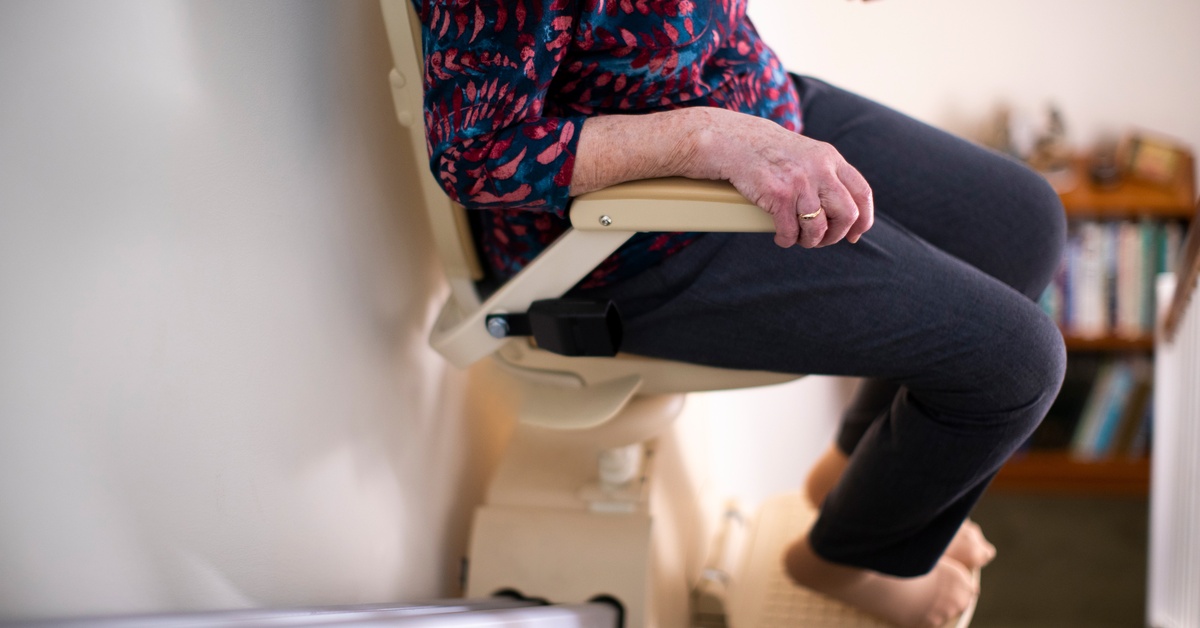 A person sits in a stairlift as the lift takes them up the flight of steps. They're wearing black pants and a floral blouse.