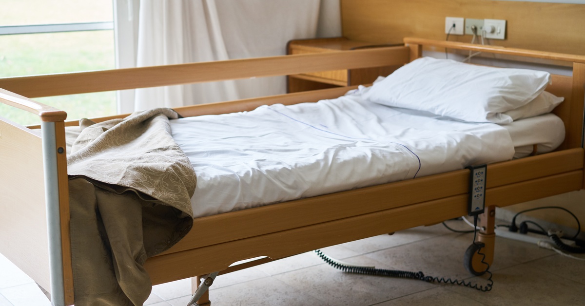 An empty rehabilitation center bed with the blanket pulled back. The sheets are white and the curtains are open.