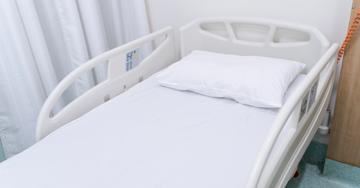 An empty hospital bed with white sheets and a bed pillow sits in an empty hospital room next to a room curtain.
