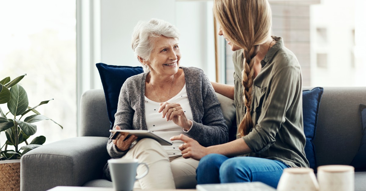 A senior woman sits on her couch smiling at her younger daughter, who sits next to her. The senior woman holds a tablet.