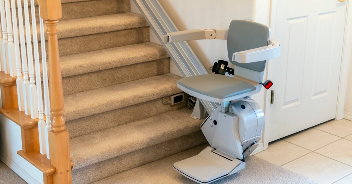 A straight stairlift parked at the bottom of a carpeted residential staircase. The banister and handrail are wooden.