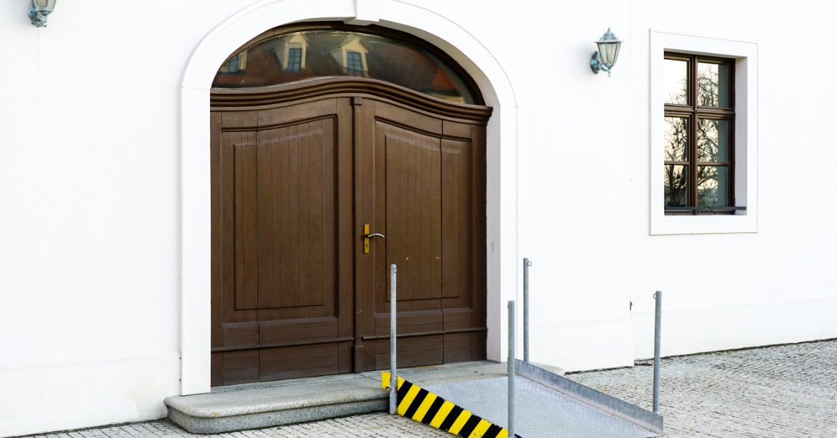 The exterior entry doors of a white church building with a stainless steel accessibility ramp placed at the door.