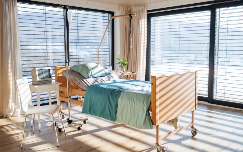 An empty hospital bed on four wheels with a wooden frame sits next to a sitting chair. The room has large windows.