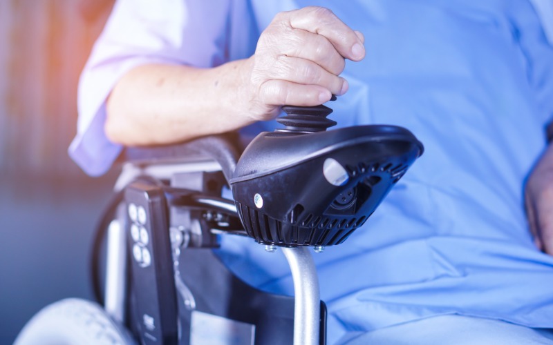 A close-up of a person riding an electric wheelchair as they use the joystick on the arm rest to navigate.