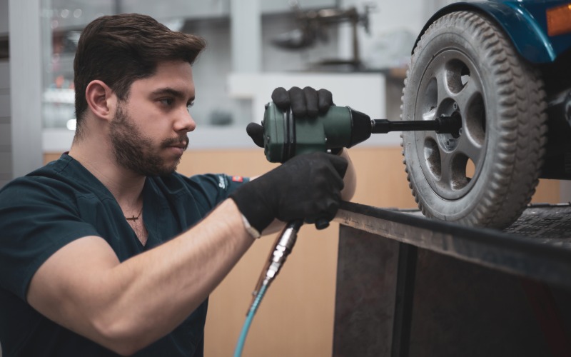 A professional wheelchair technician takes a drill to the wheelchair wheel. He wears black scrubs to work.