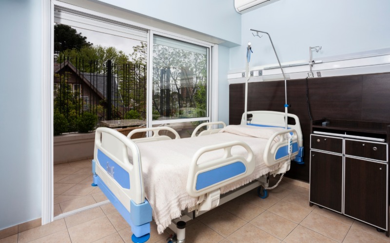 A hospital bed rental sits next to the sliding glass door in the rehab facility room. The linens are white.