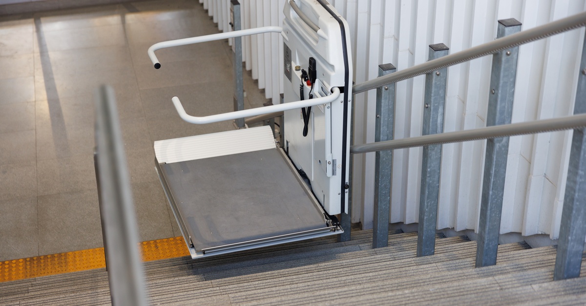 A stairlift at the bottom of a stairwell. The platform and the arm rests are down and the lift is in a covered stair area.