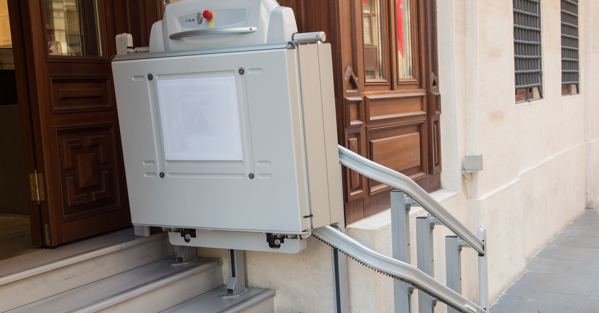 An outdoor stairlift on the side of a building at the top of the stairs. The seat is folded up, and there's a red button.