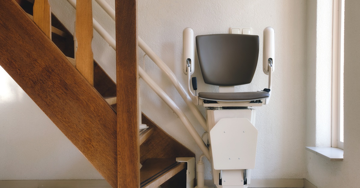A home stair lift sits at the bottom of the staircase. The staircase has a wooden handrail and wooden steps.