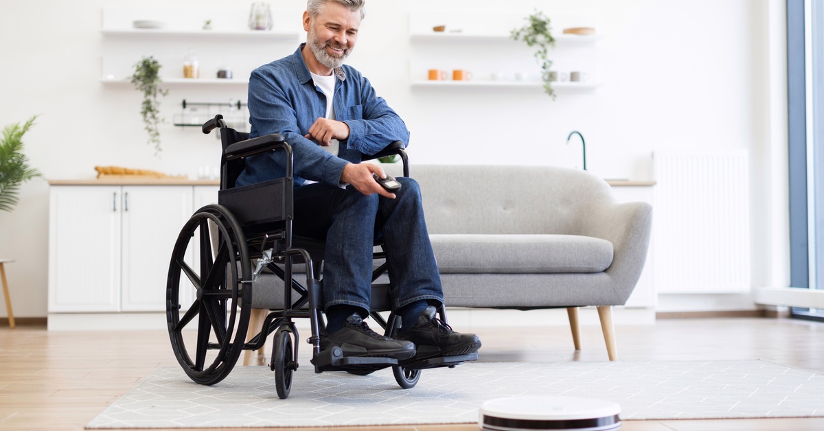 A man at home in a wheelchair watches the robot vacuum glide across his living room floor. There's a white couch behind him.