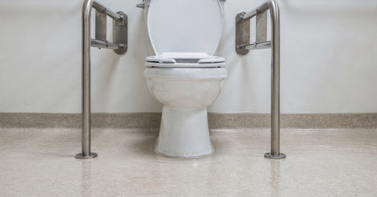 A long-term care facility with a toilet on in the bathroom surrounded by stainless steel accessibility grab bars.