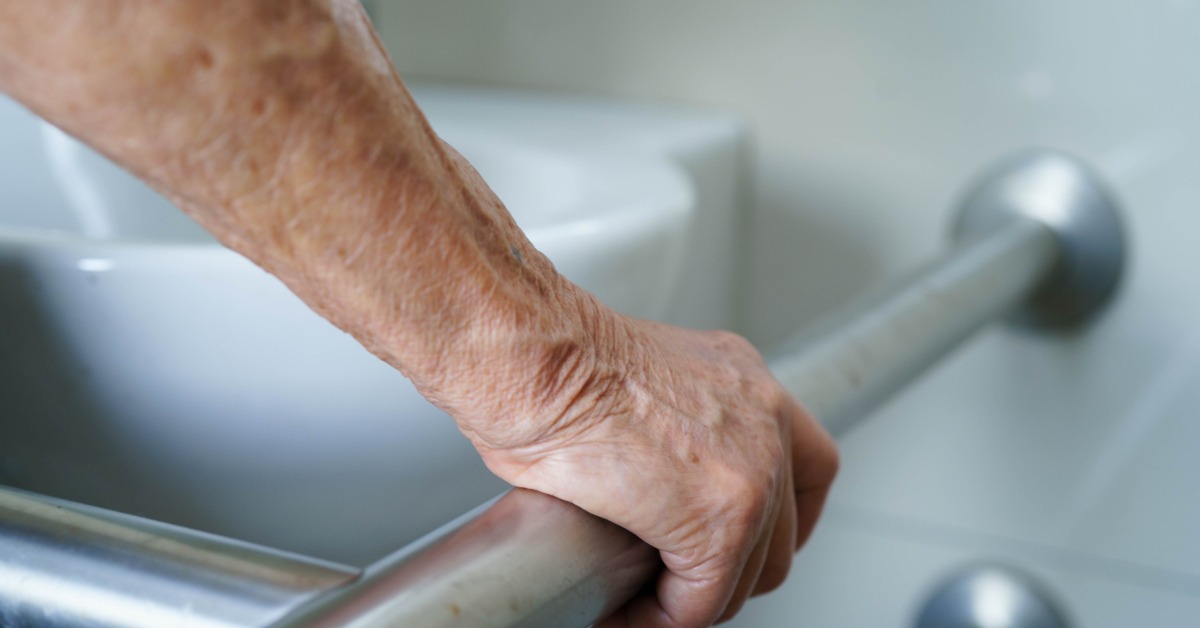 A close-up of an older adult's arm grabbing a grab bar in the bathroom to use the toilet. The bar is installed to the wall.